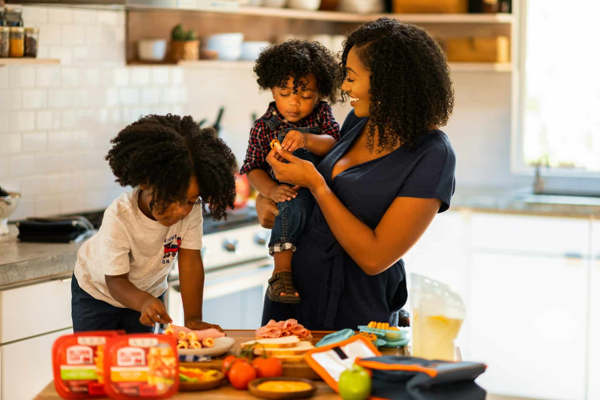 A smiling Black mother holds a toddler while another young child stands at a kitchen counter preparing lunch items.