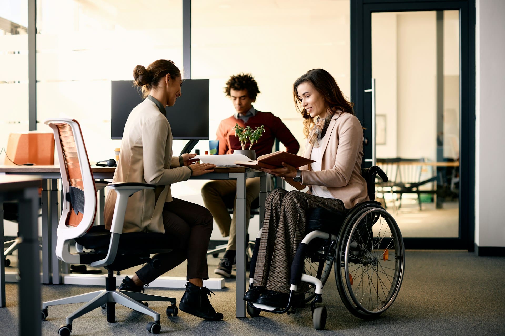 A woman in a wheelchair collaborates with her co-workers, a man and a woman, around a desk in an office, representing disability in employment.