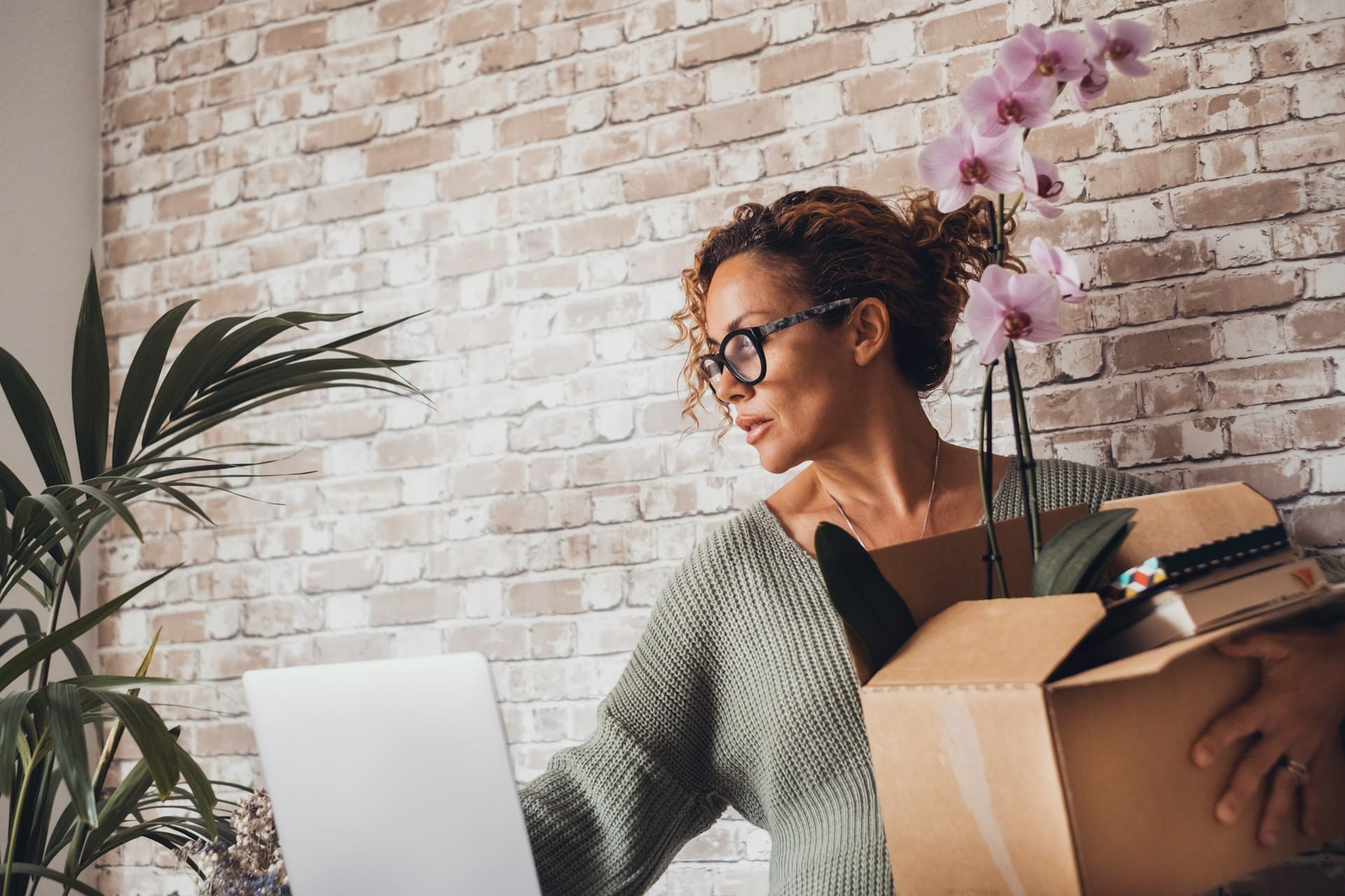 A recently laid off woman carries a cardboard box with her office belongings in one hand, while reaching for her laptop with the other, representing employees who have been laid off.