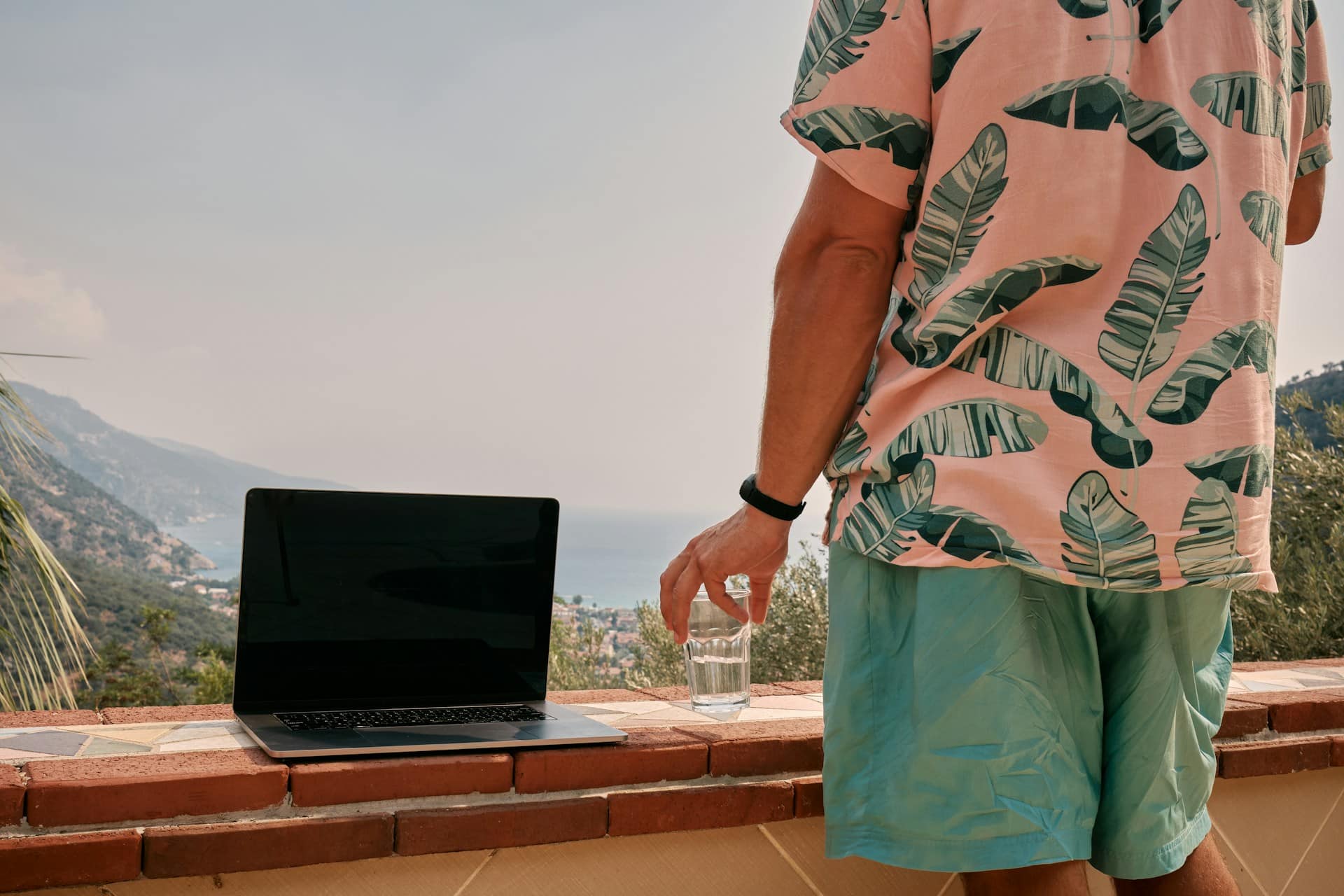 A man holding a cocktail and looking out over a tropical vista with his laptop sitting on a platform next to him, representing unauthorized remote work and