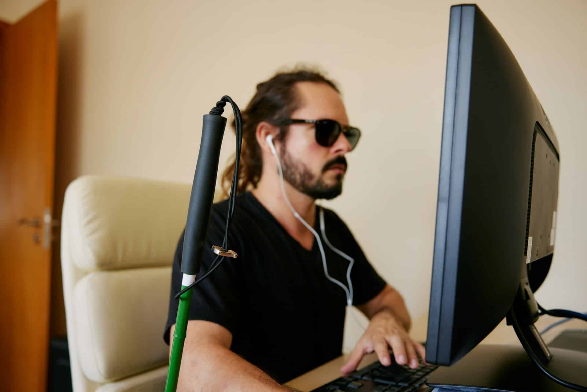 A blind man sits at a desk in front of a computer screen. He is wearing subglasses and earbuds, and his cane leans against the desk, representing disability accommodations in hiring.