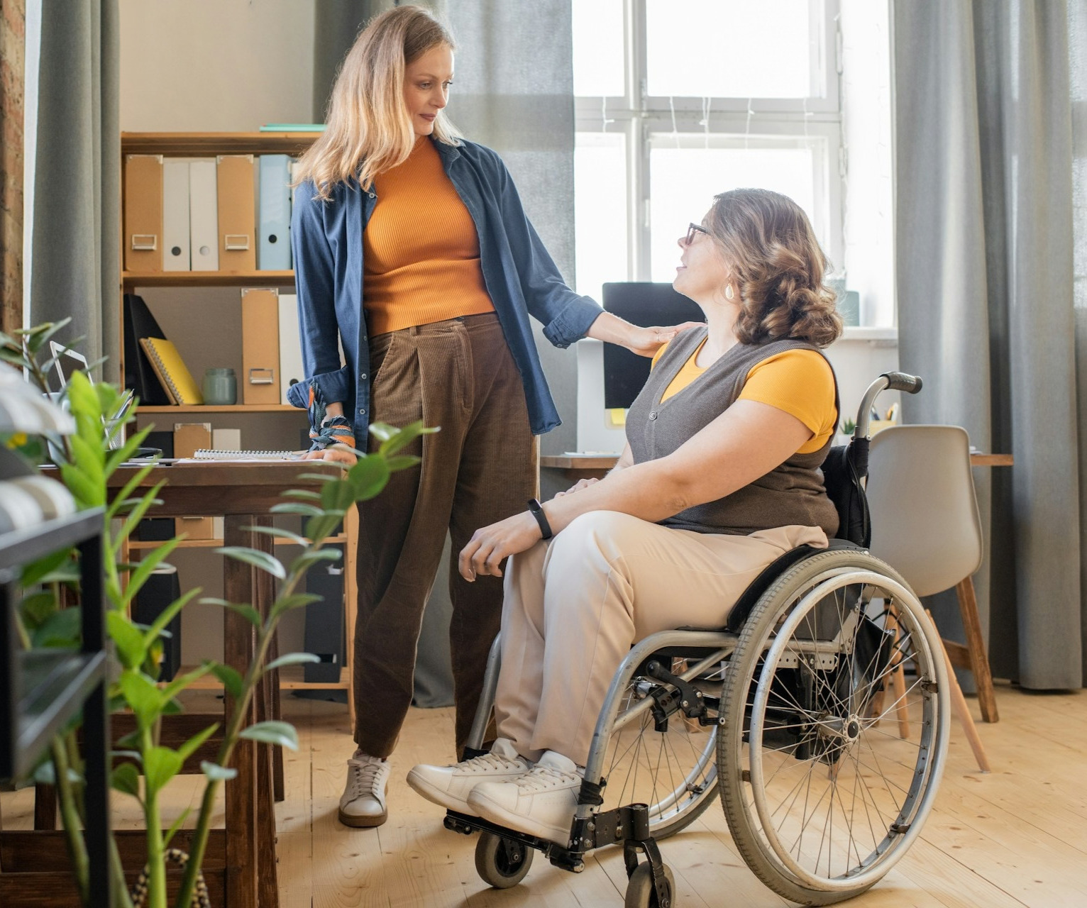 In an office a woman rests her hand on a woman in a wheelchair's shoulder, representing workplace accommodation plans.