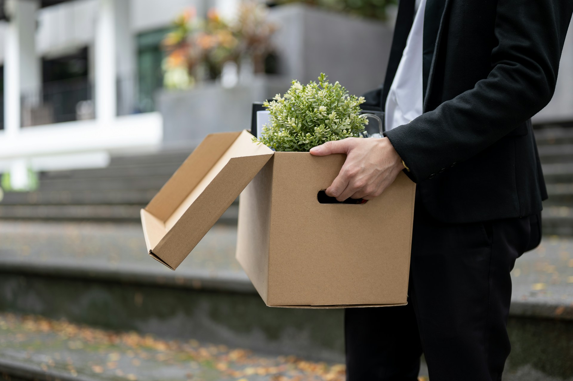 A man, shown from the shoulders down, carries a box of belongings from his office after being terminated, representing RSUs and termination pay.