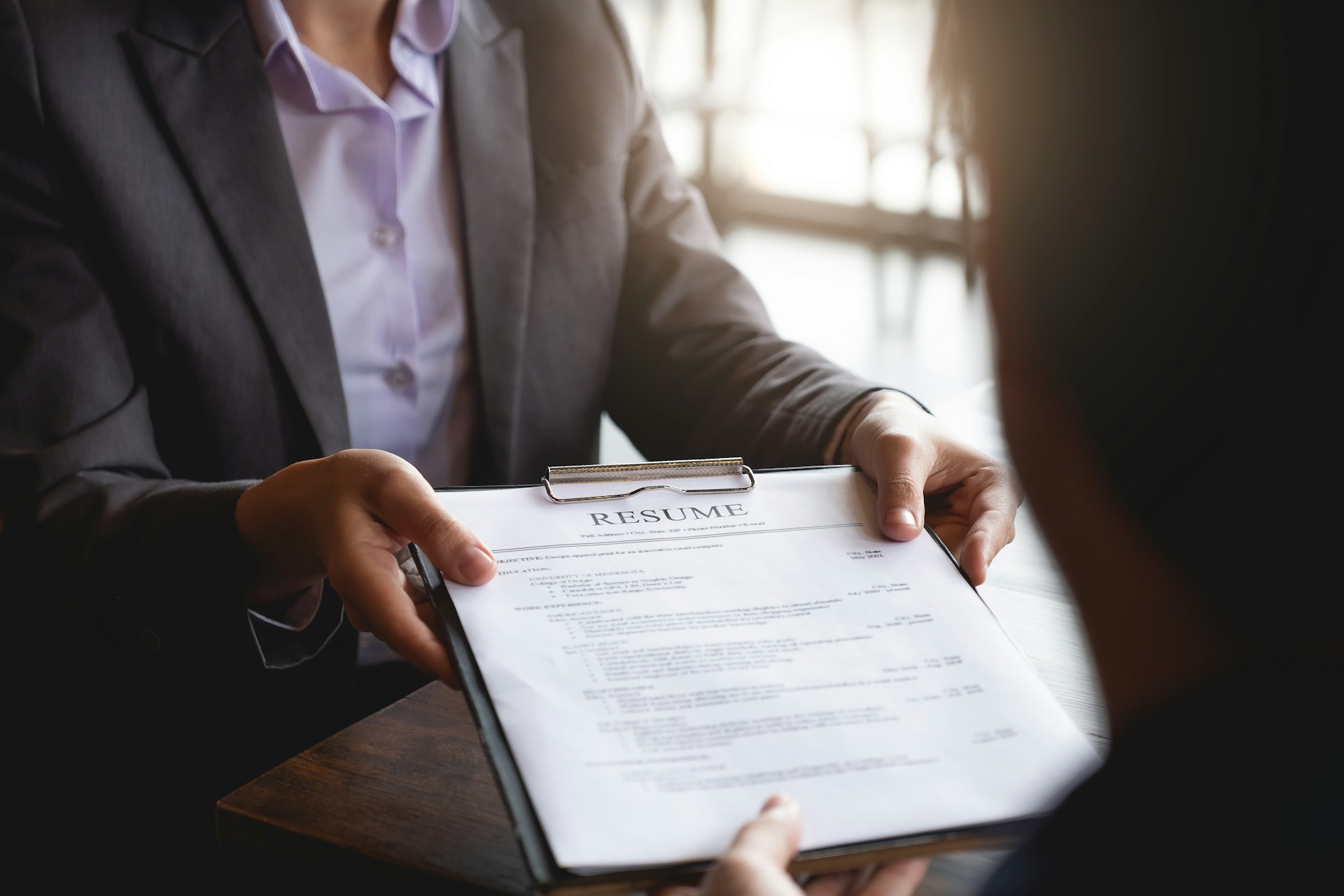 A person holding a clipboard with a printed copy of a resume handing it to another person. This photo could represent looking for another job while already employed.
